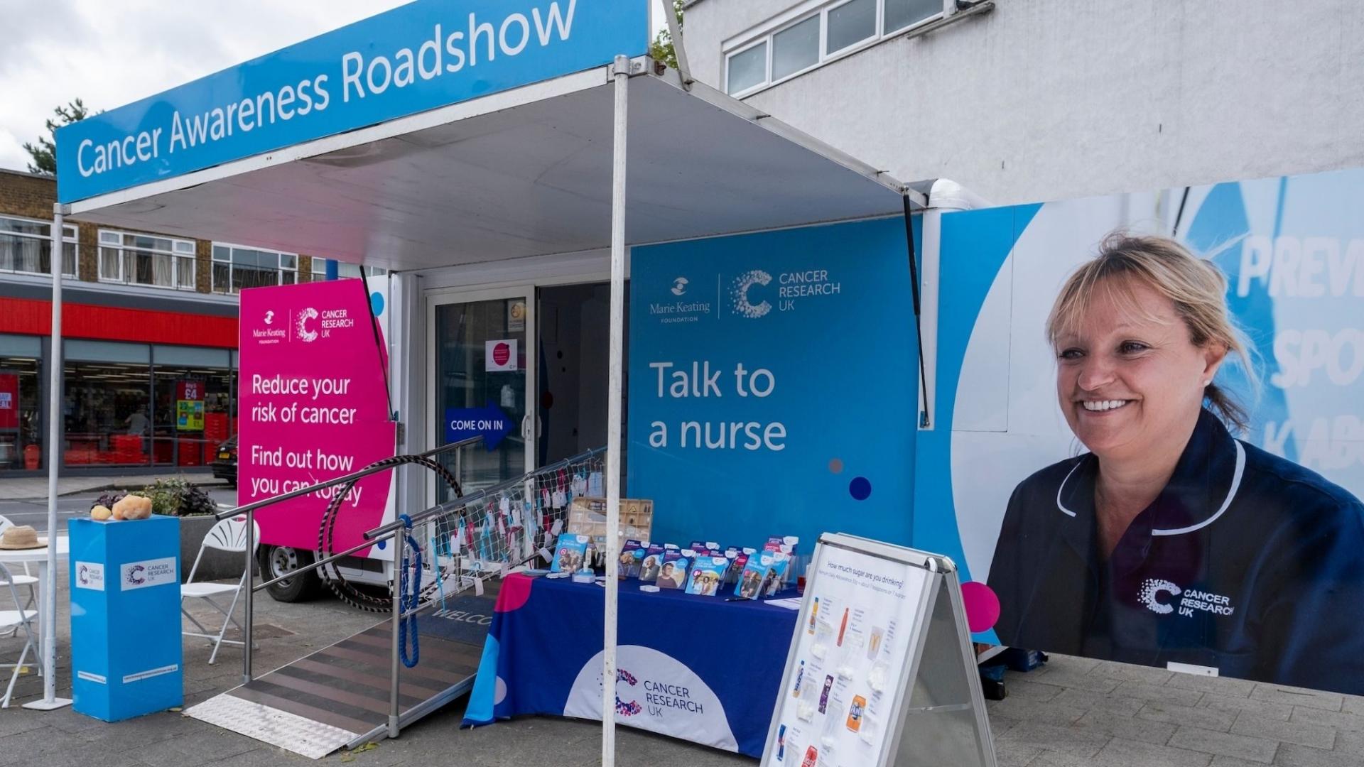 Photo of a blue and white Cancer Research UK Roadshow truck parked outside with a table and pop up stand set up with informative materials