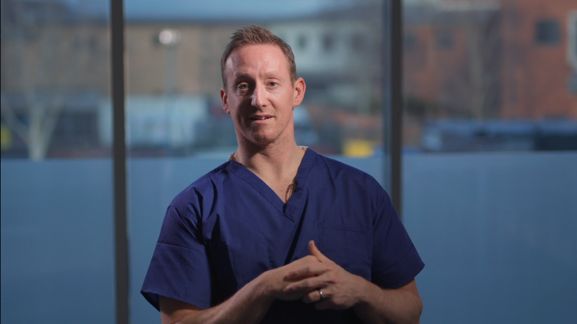 Photo of Dr Matt Evison wearing blue scrubs sitting in front of a window overlooking a river