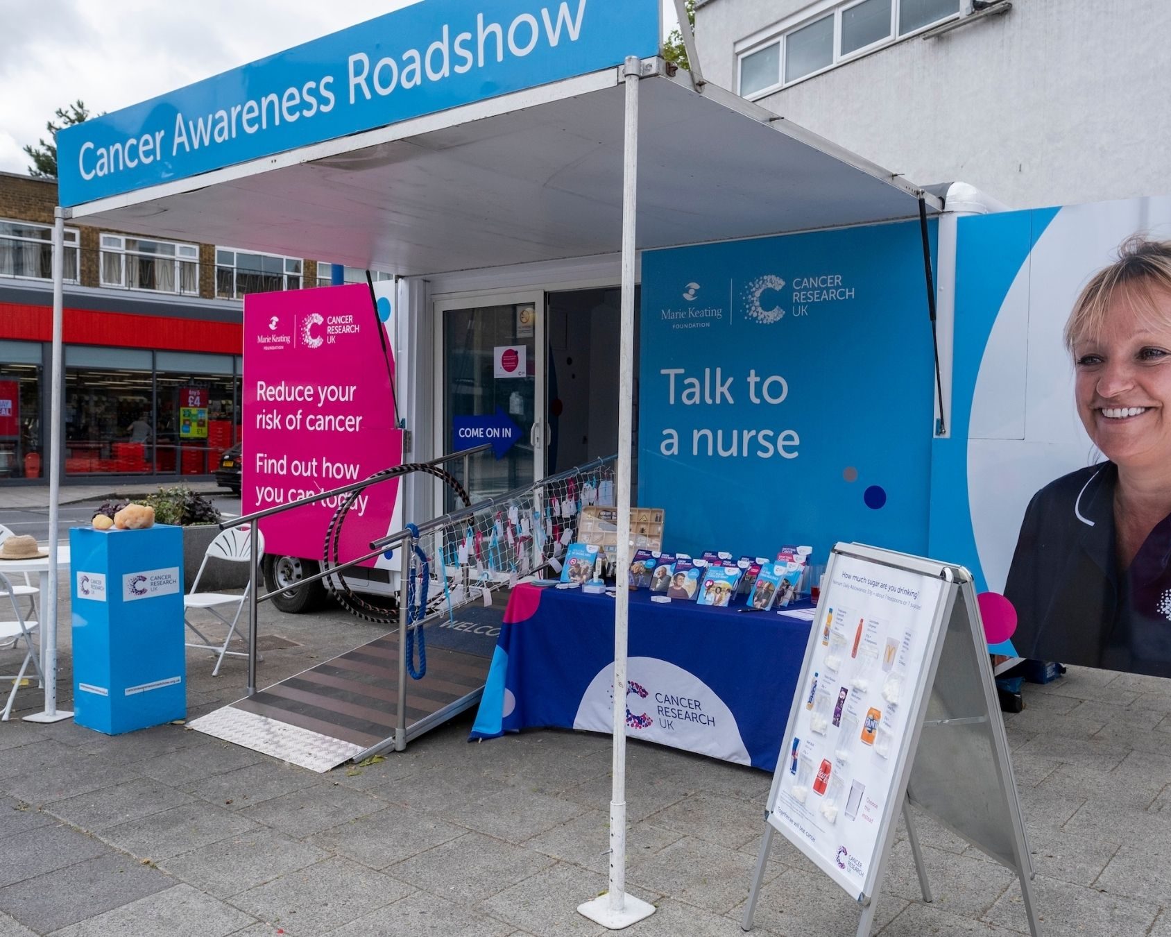 Photo of a blue and white Cancer Research UK Roadshow truck parked outside with a table and pop up stand set up with informative materials