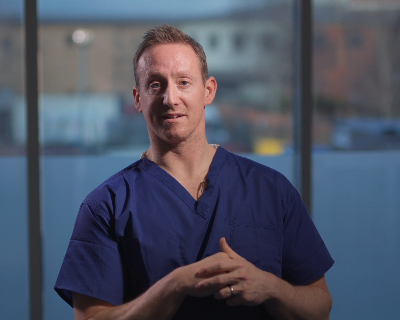 Photo of Dr Matt Evison wearing blue scrubs sitting in front of a window overlooking a river